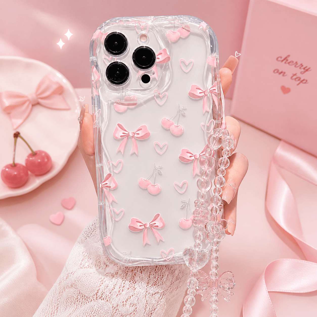 Clear iPhone case with wavy scalloped edges, covered in raised pink bows, pink cherries and small hearts, with a clear beaded wrist strap and heart charm attached