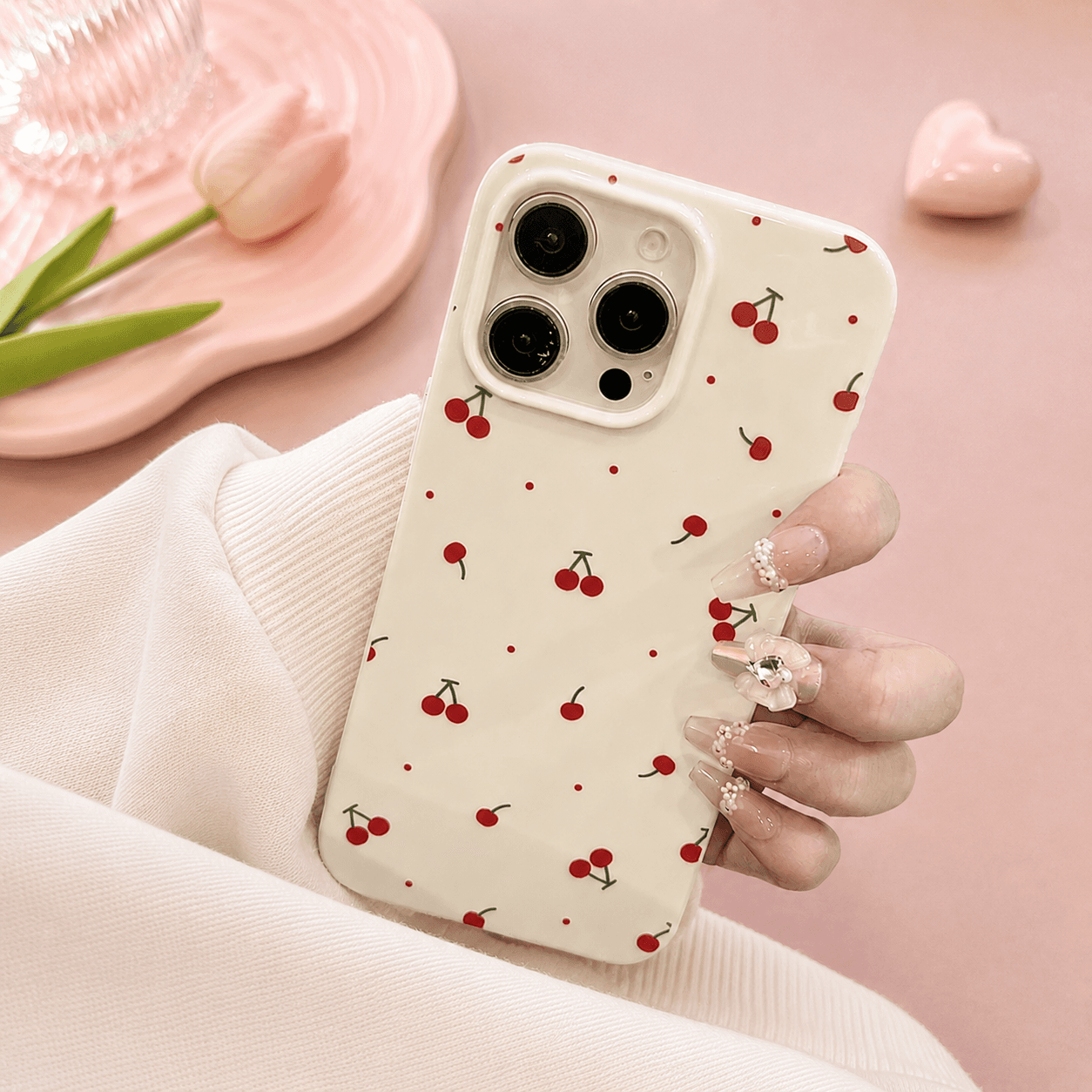 Cream iPhone case printed with small red cherries on green stems and scattered red dots, held in a hand against a pink background